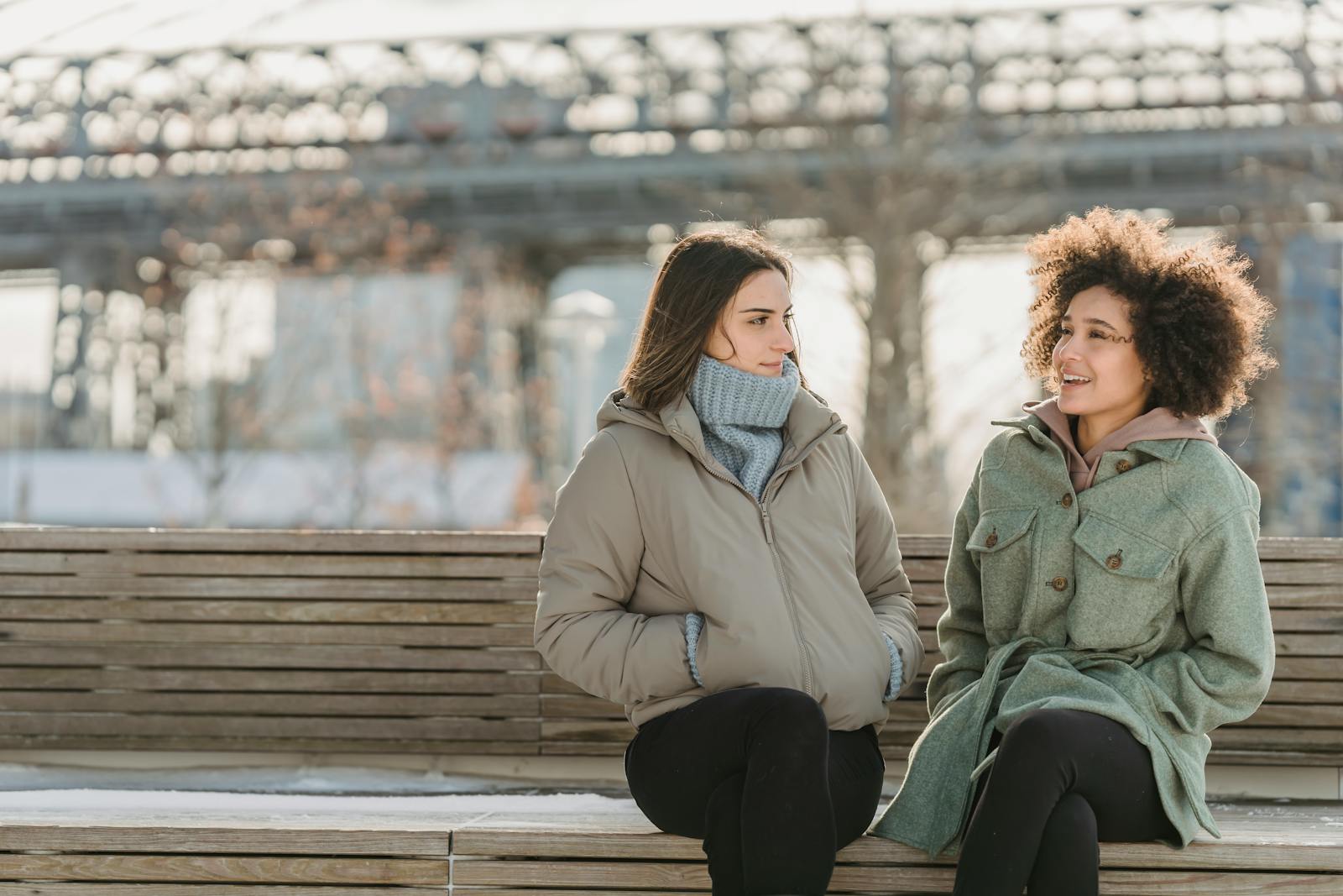 Cheerful multiracial female friends in warm clothes sitting on wooden bench while spending time together on street in winter day