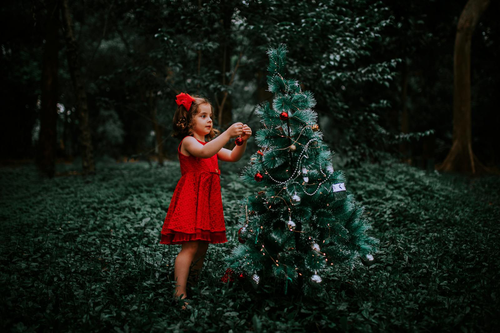 Young girl in a red dress decorates a Christmas tree outdoors, embodying festive spirit.