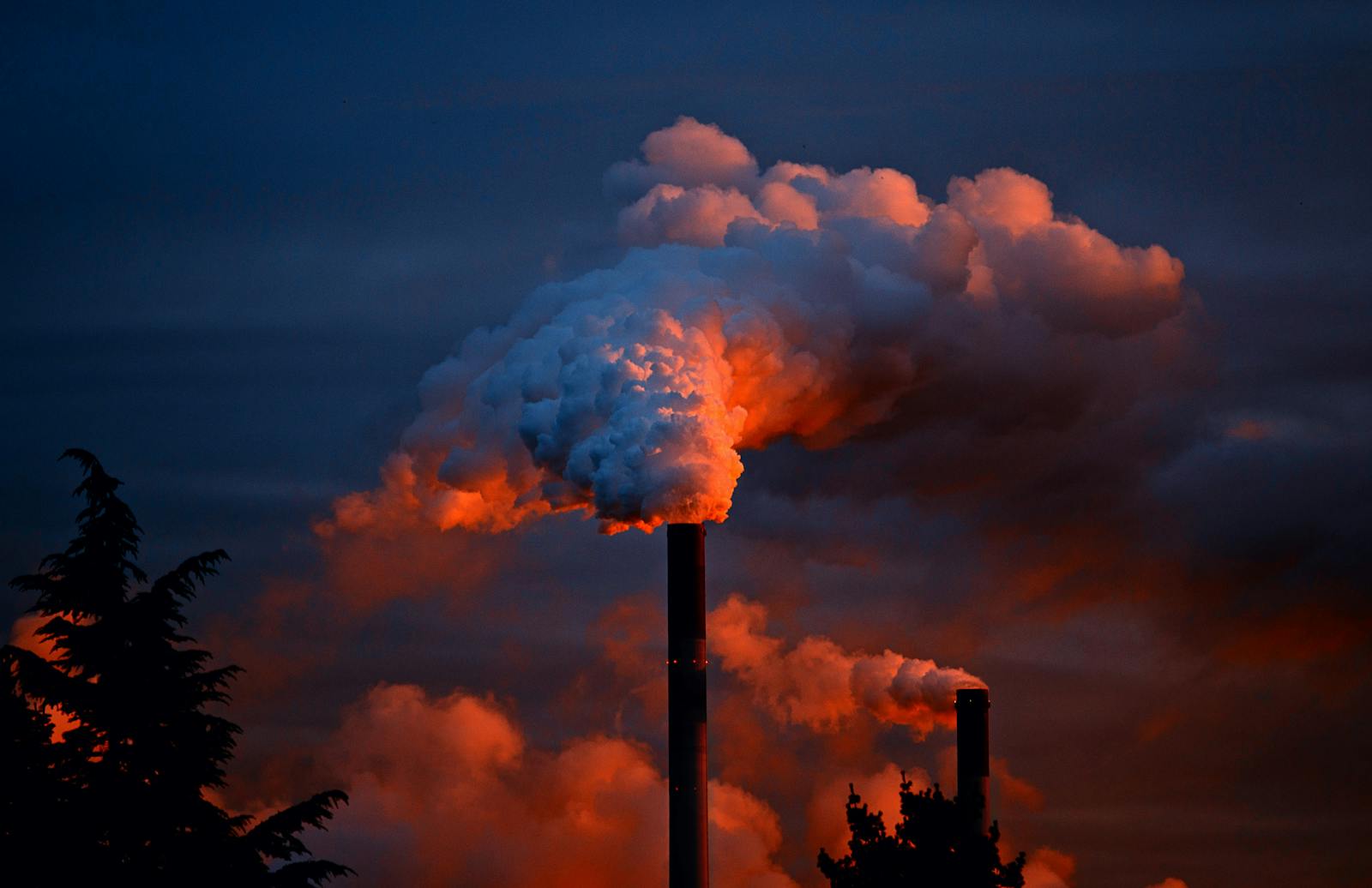 Smoke billows from industrial chimneys at sunset, highlighting pollution against a vibrant sky.