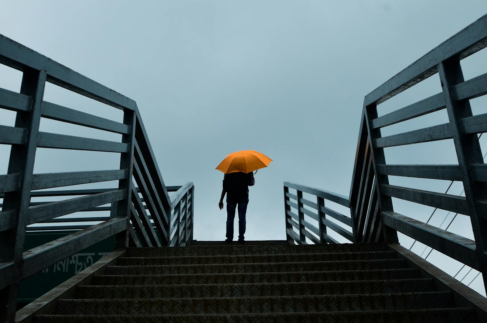Silhouette of a person holding an orange umbrella on a steel footbridge, low angle view.
