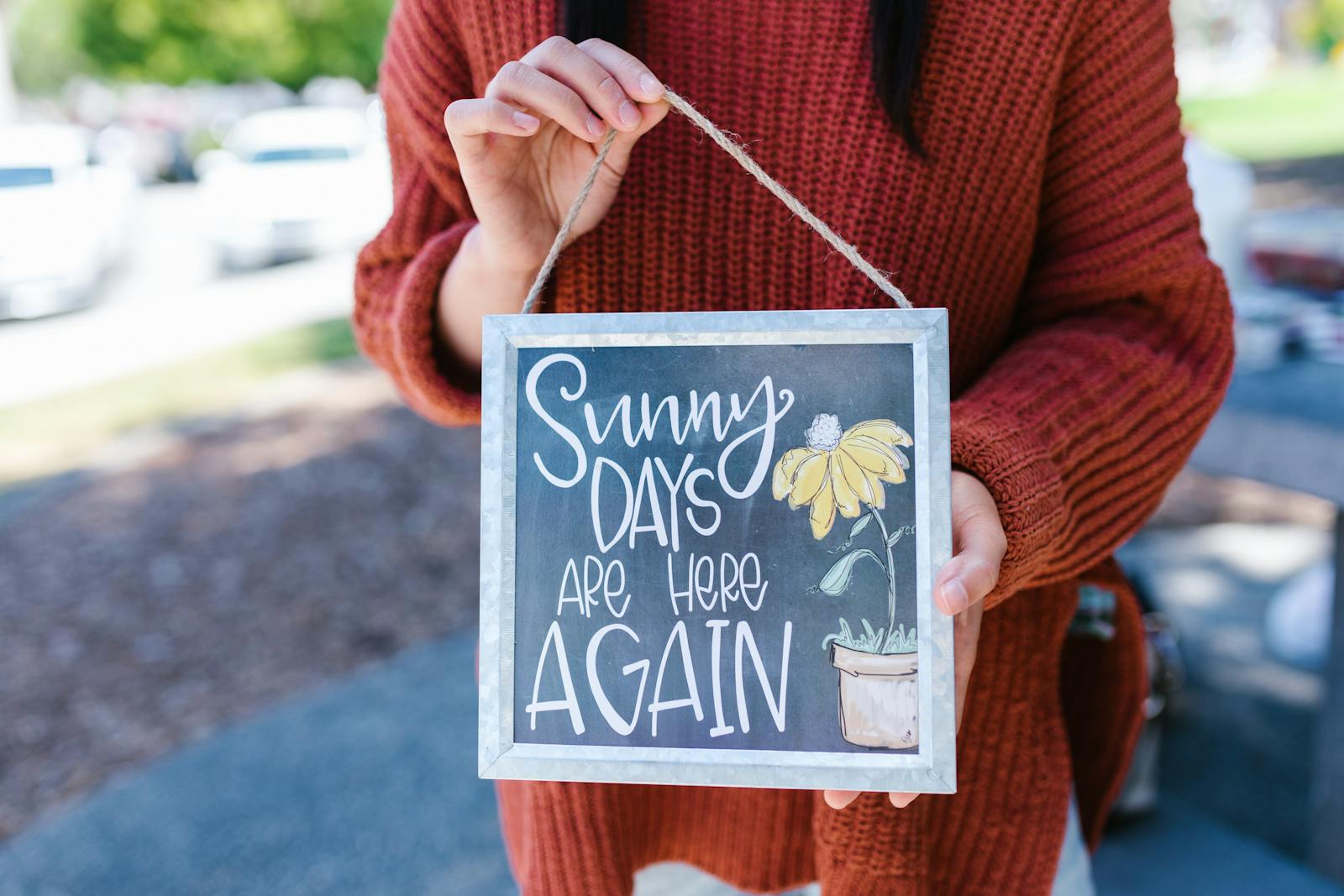 Woman in a sweater holding a sign with 'Sunny Days Are Here Again' text outdoors.