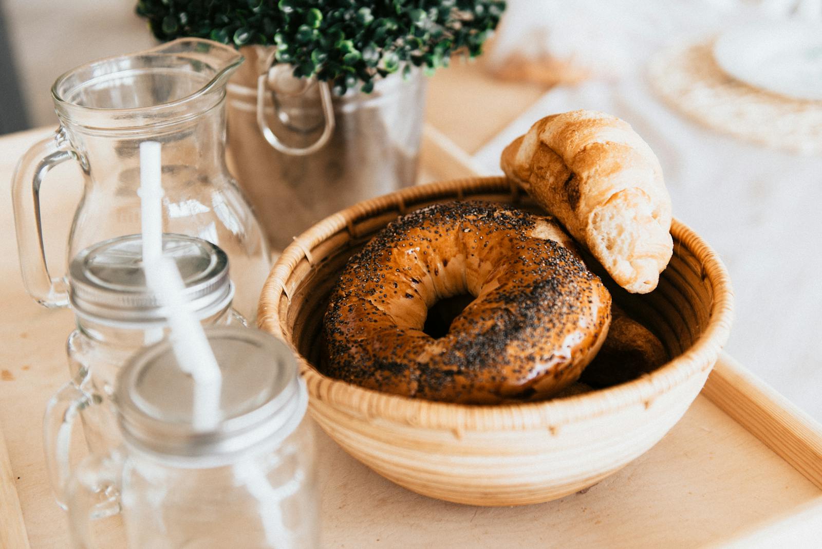 Fresh bagels and croissant on a wooden tray with mason jars, creating a warm rustic breakfast vibe.