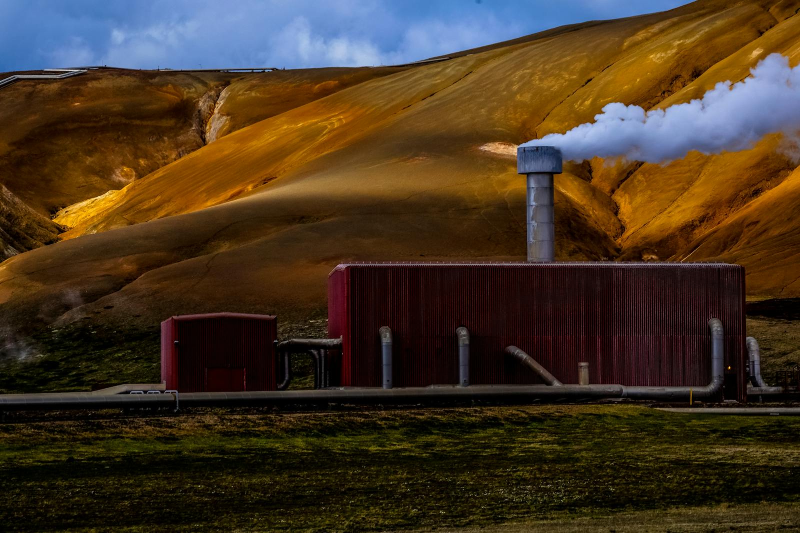 Red geothermal power plant emitting steam against a dramatic yellow-brown mountainous backdrop.