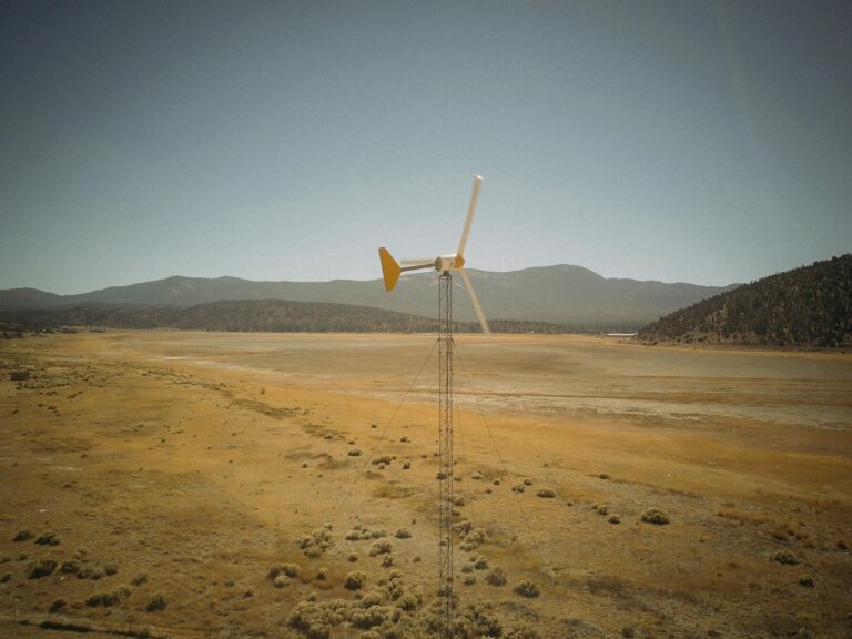 A solitary wind turbine standing in a barren desert landscape with mountains in the background.