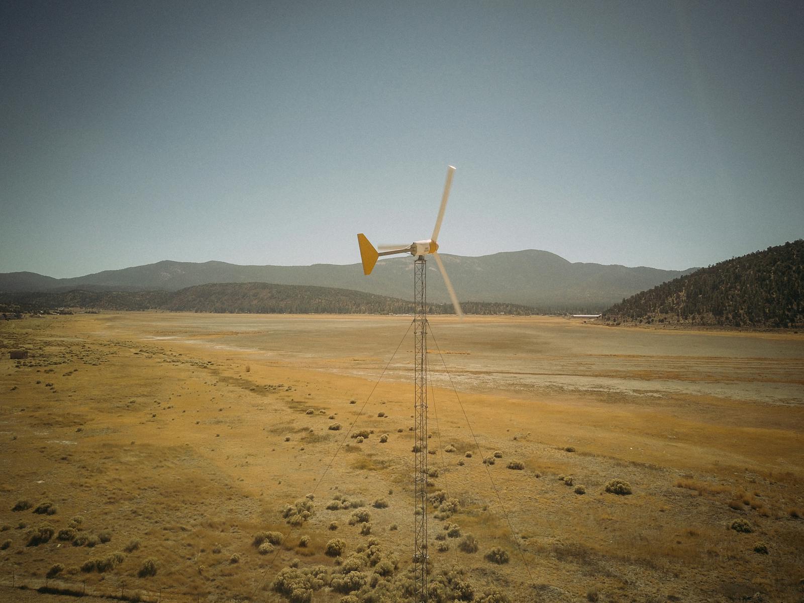 A solitary wind turbine standing in a barren desert landscape with mountains in the background.