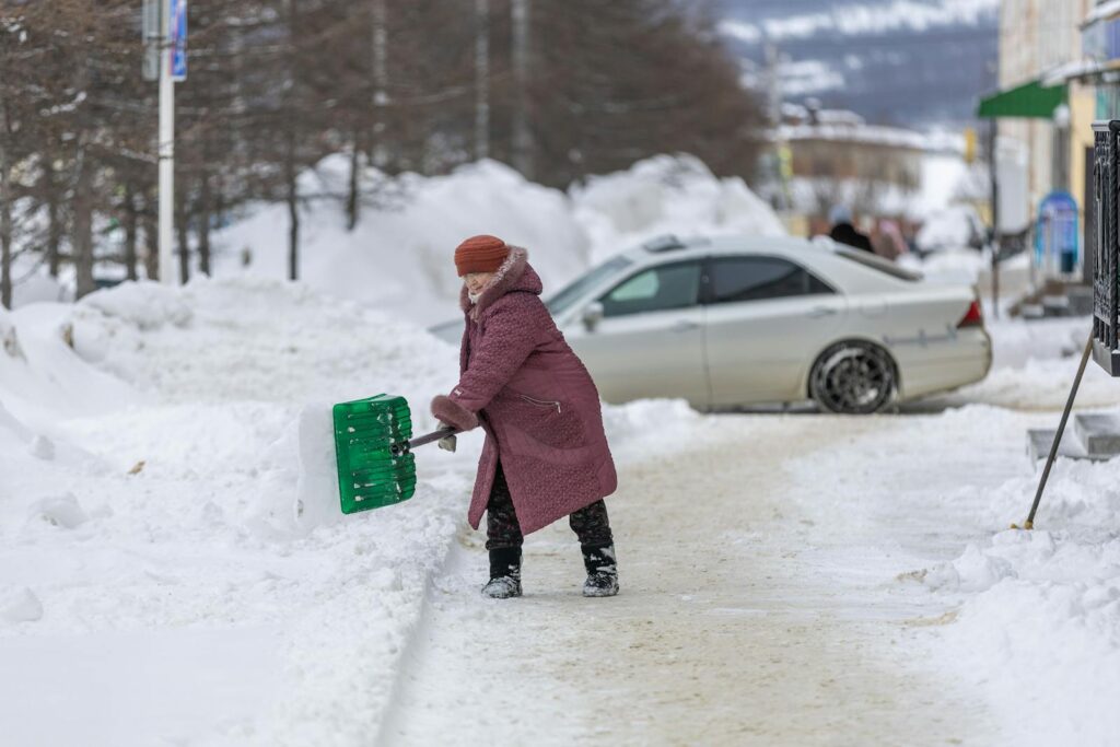 Elderly woman in a pink coat shoveling snow on a street, capturing the essence of winter chores.