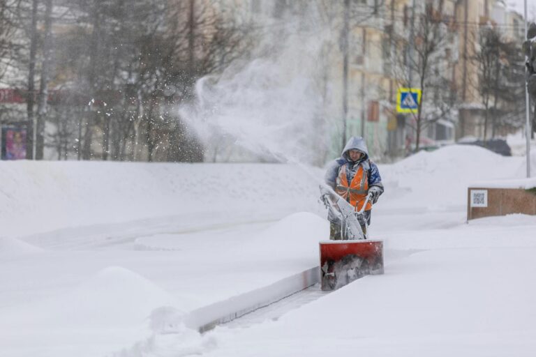 Person using a snow blower to clear a city sidewalk during a heavy winter snowfall.
