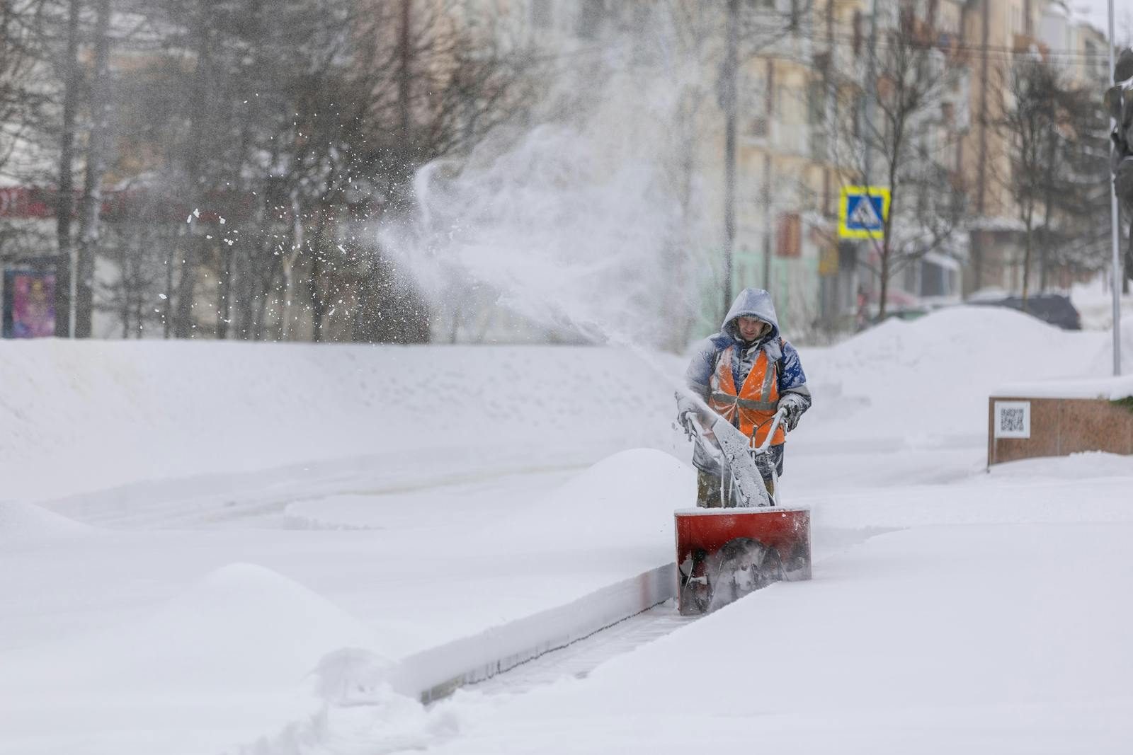 Person using a snow blower to clear a city sidewalk during a heavy winter snowfall.