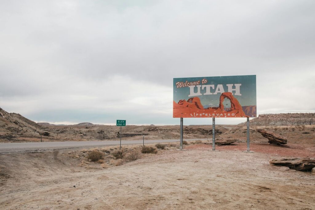 A 'Welcome to Utah' sign with desert landscape background under a cloudy sky.