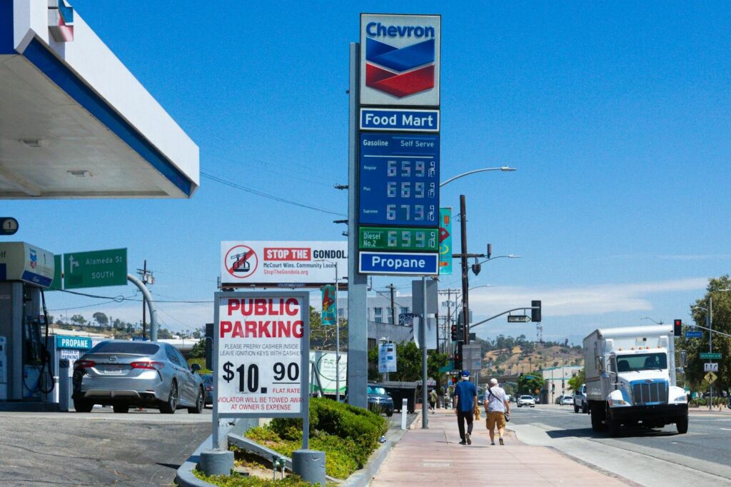 People walking near a Chevron gas station with prominent signage and parked vehicles on a sunny day.