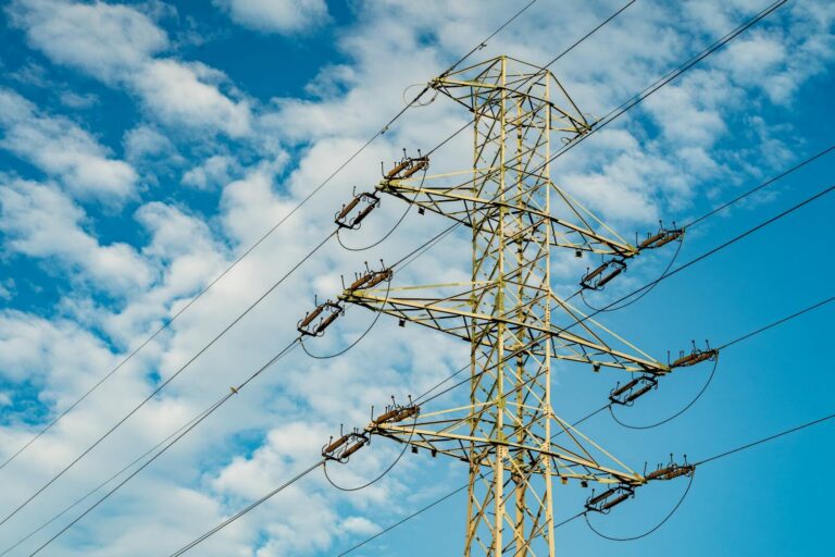 A tall electrical transmission tower stands against a backdrop of blue sky and white clouds.