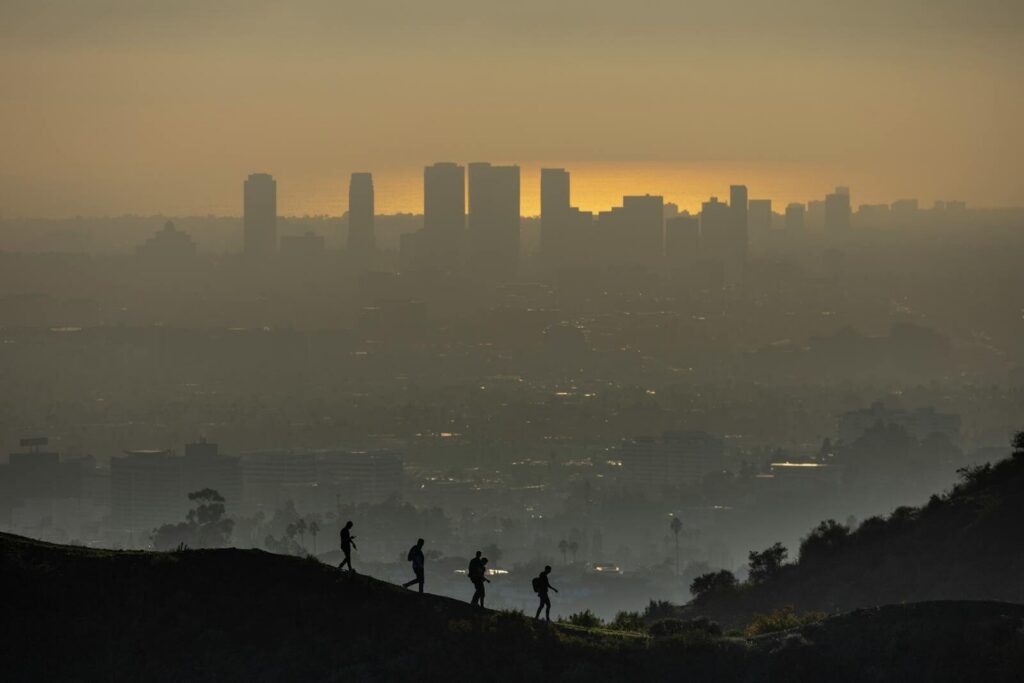 Silhouetted hikers traverse a hill, overlooking the smoggy Los Angeles skyline at sunset.