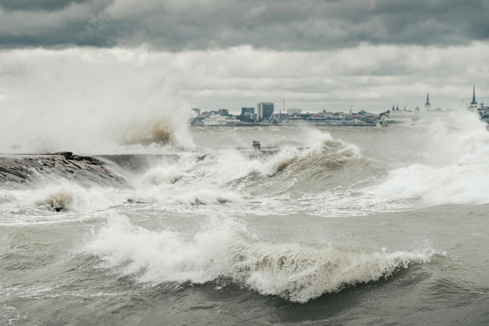 Powerful ocean waves crashing against a rocky coastline under a stormy sky with distant city skyline.
