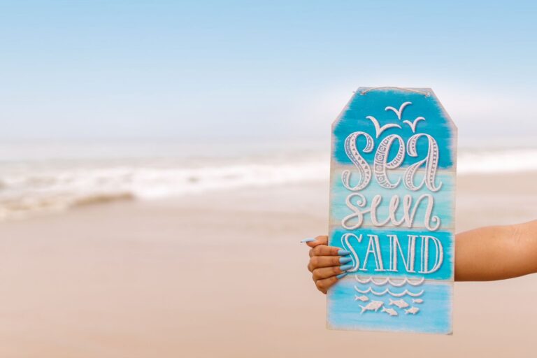 Hand holding a sign with 'Sea Sun Sand' against a tropical beach background.