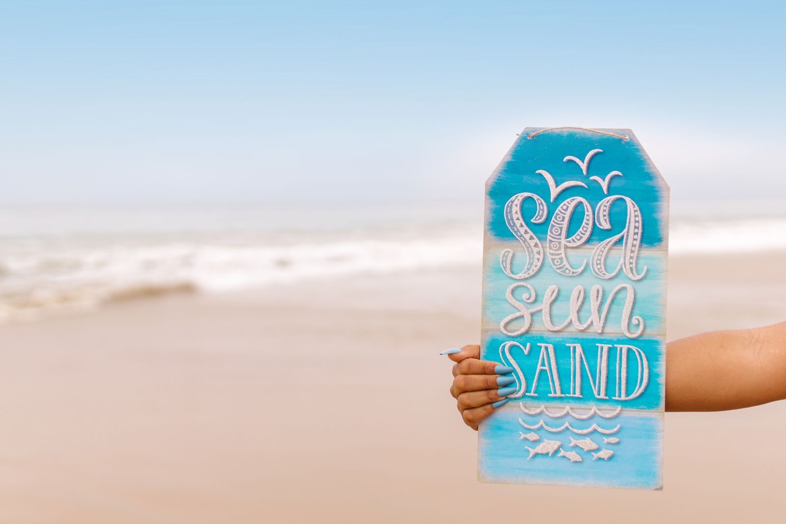 Hand holding a sign with 'Sea Sun Sand' against a tropical beach background.