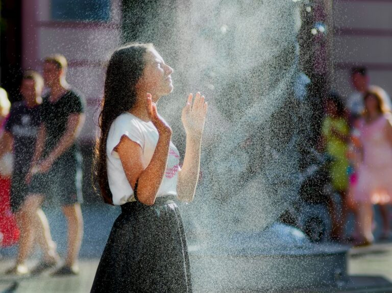 A young woman stands in refreshing mist outdoors, enjoying the cool water spray on a sunny day.