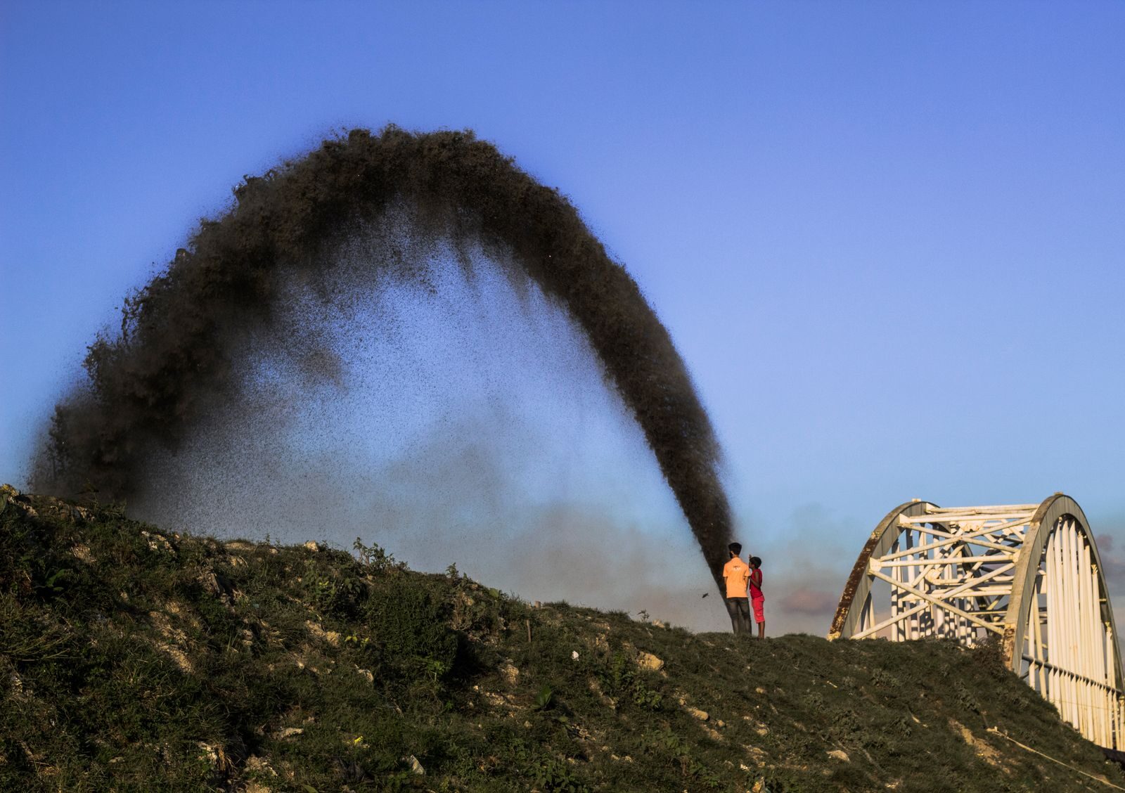 Industrial waste spills over a hill near a bridge, highlighting environmental impact.