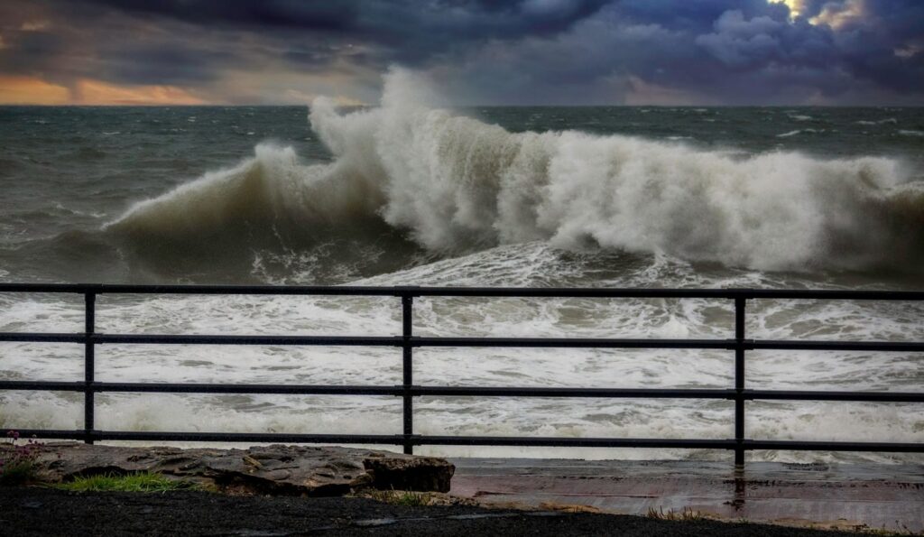 Powerful ocean waves crash against a pier with dramatic skies overhead.