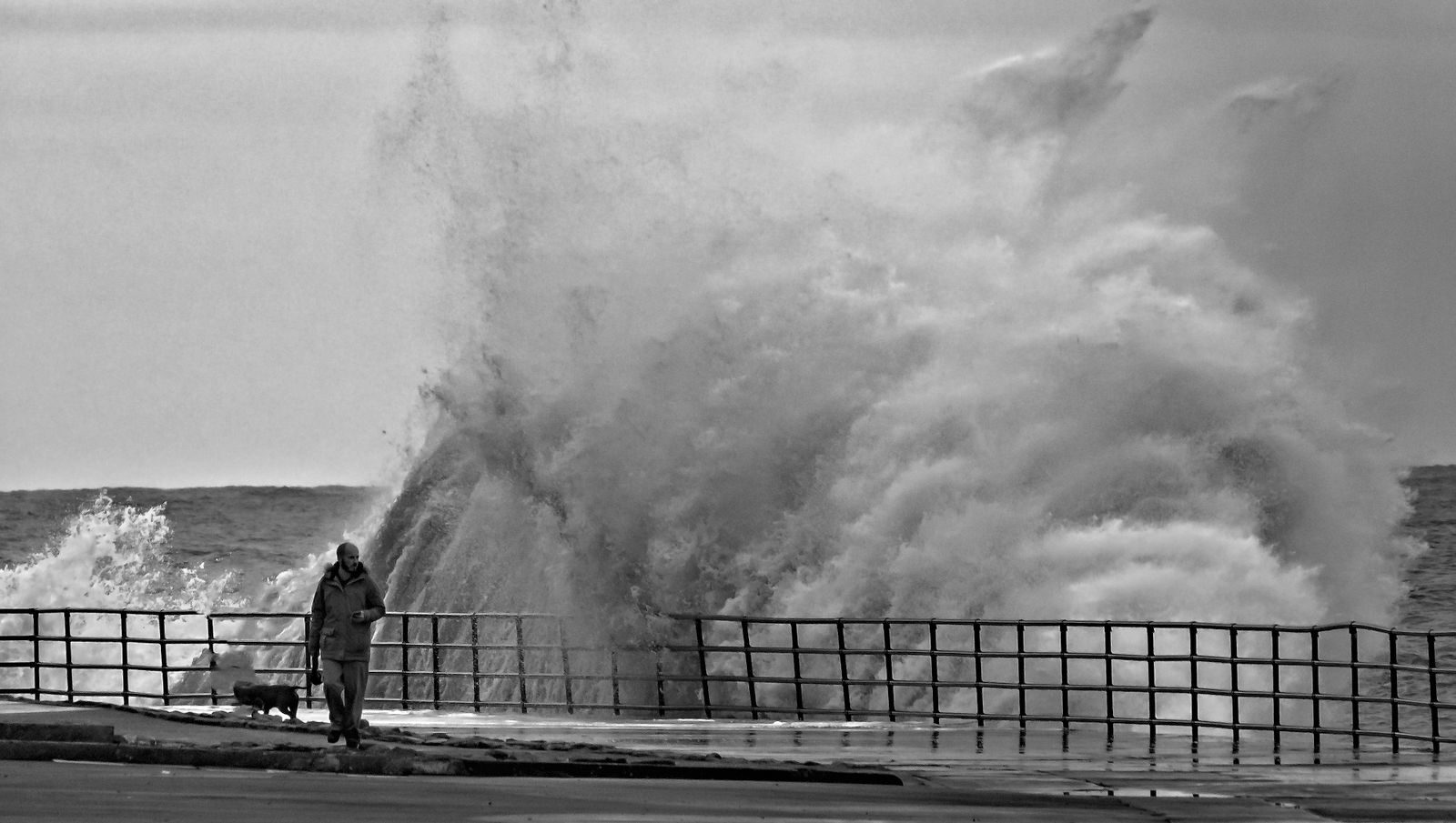 Powerful storm wave crashing against a dock with a solitary person braving the stormy sea.