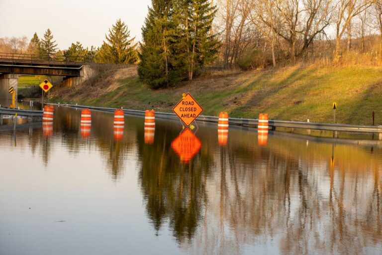 A flooded road with warning signs indicating 'Road Closed Ahead' due to water overflow.